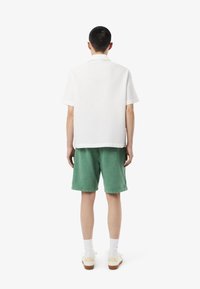 Chemise blanche texturée à manches courtes associée à un short vert ample, complété par des chaussettes blanches et des baskets beiges avec des semelles marron.