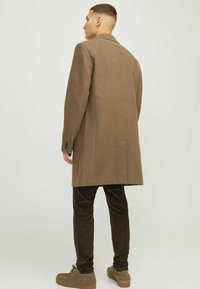 Abrigo de mezcla de lana marrón con silueta recta, solapas con muesca y una abertura trasera. Combinado con pantalones de pana oscuros y zapatos marrones.