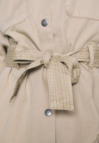 Beige jakke med bælte, sølvfarvede trykknapper, et tekstureret bælte med syninger og store forlommer. Blødt, holdbart stof.