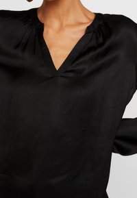 Blusa nera in satin con scollatura a V, tessuto arricciato sulle spalle e superficie liscia. Maniche lunghe con vestibilità rilassata.