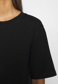 Henkilö, jolla on päällä yksinkertainen musta lyhythihainen crew neck -t-paita tasaisella valkoisella taustalla, näkyvissä kaula, hartia ja yläkäsivarsi.