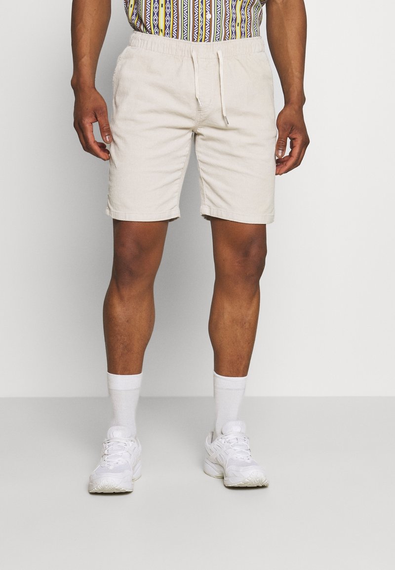 Shorts en coton beige avec une taille élastique et un cordon de serrage, dotés d'une coupe droite et d'ourlets retroussés. Portés avec des baskets blanches et des chaussettes.