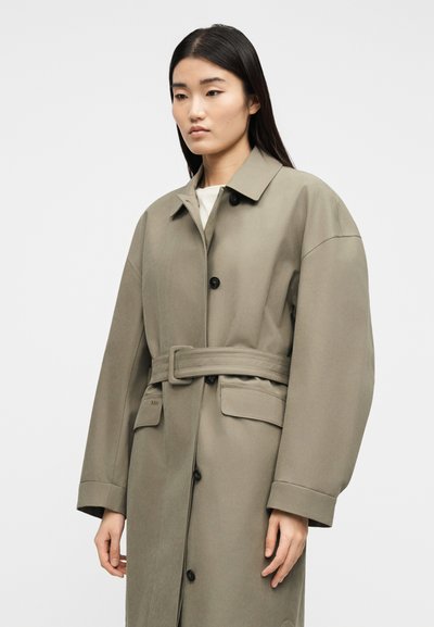 Γυναίκα με μακριά σκούρα μαλλιά που φοράει ένα ζαρωτό, φαρδύ παλτό τυπου τρανς σε ταφ χρώμα με μαύρα κουμπιά και τσέπες με καπάκι, μπροστά σε απλό φόντο.