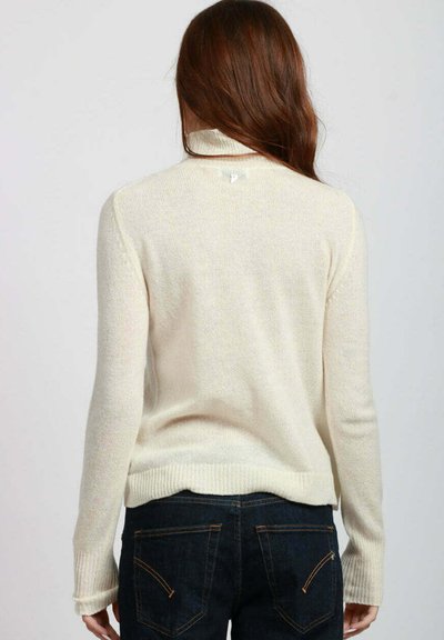 Donna con lunghi capelli castani che indossa un maglione a collo alto color crema e jeans blu scuro, vista di spalle su uno sfondo semplice.