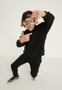 Melna kapuce un sporta bikses komplekts ar priekšējo kabatu. Aksesuāri ietver melnu cepuri un gaiši rozā atstarojošas saulesbrilles. Melni un balti sporta apavi.