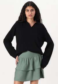 Svart genser med lange ermer og V-hals samt en avslappet passform, sammen med et grønt, lagdelt rynket skjørt. Glatt tekstur i stoffet gjennomgående.