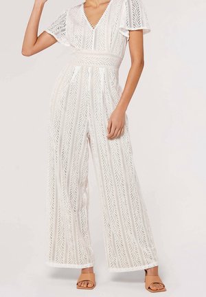 Kobieta w szerokim kombinezonie z białej koronki z rozkloszowanymi rękawami i głębokim dekoltem w serek, w połączeniu z brązowymi sandałami na słupku z odkrytymi palcami.