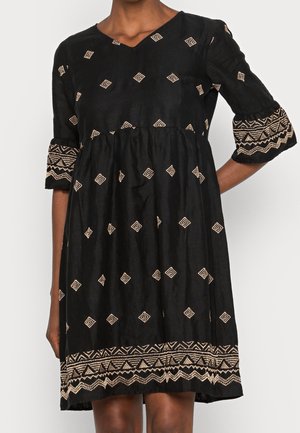 Kvinde iført en sort knælængde kjole med beige geometrisk broderi på ærmerne, kanten og spredte diamantmønstre.