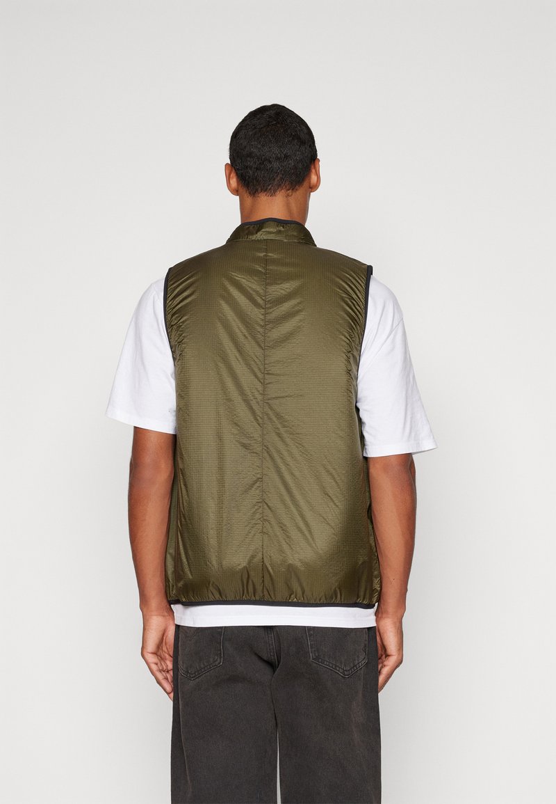 Gilet matelassé vert olive avec un col montant, texture matelassée et fermeture éclair à l'avant. Porté par-dessus un t-shirt blanc et un pantalon noir.