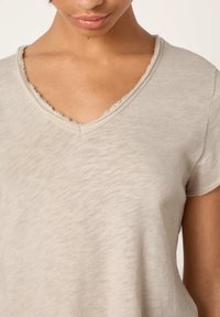 Žena v béžovom tričku s výstrihom do V, s ošúchanými okrajmi, ukazujúca hornú časť hrudníka, krk a čiastočne tvár.
