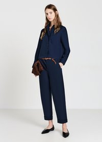 Marineblaue Langarmbluse mit Knöpfen und Brusttasche, kombiniert mit marineblauen Hosen mit umgeschlagenem Saum. Das Modell hält eine braune Wildleder-Clutch und trägt schwarze Ballerinas.