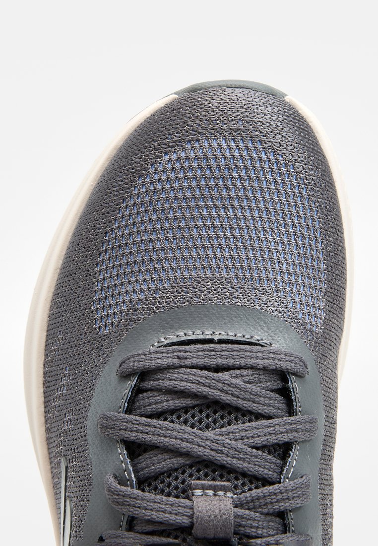 Zapatilla deportiva gris con parte superior de malla texturizada y acentos azules, cuello acolchado y cordones planos. La suela de goma es blanca.