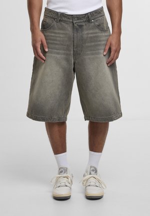 Muž oblečený v vybledlých sivých riflových krátkych nohaviciach po kolená, bielych ponožkách a béžových teniskách New Balance, stojaci pred jednoduchým pozadím.