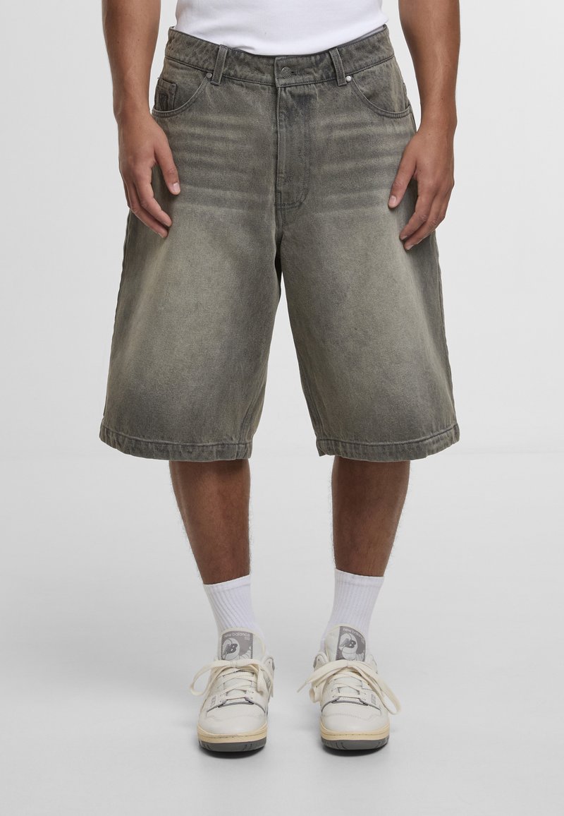 Mees, kes kannab tuhmunud halli põlvikutega teksapükse, valgeid sokke ja beeže New Balance'i tossusid, seisab ühetaustalise tausta ees.