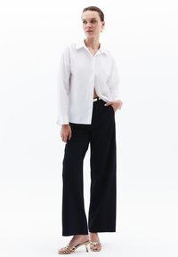 Witte blouse met lange mouwen en kraag, gecombineerd met hooggemiddelde, wijde zwarte broek en gouden sandalen met bandjes.