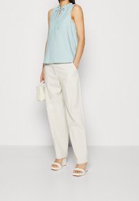 Top sin mangas de color azul claro con cuello volante y detalle de lazo, combinado con pantalones anchos blancos y sandalias blancas. Sosteniendo un pequeño bolso beige.
