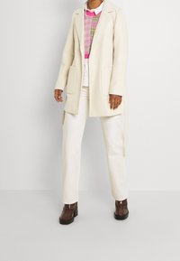 Cappotto lungo color crema con una vestibilità sartoriale, grandi tasche, indossato sopra un maglione a fantasia rosa e verde, abbinato a pantaloni bianchi e tacchi a piattaforma marroni.