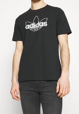Mężczyzna w czarnej koszulce Adidas z białym logo i czarnych dżinsach, stojący na tle jednolitego jasnego tła.
