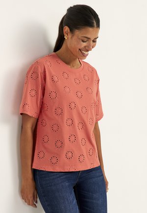 Vrouw met donker haar in een paardenstaart, draagt gouden oorbellen, een koraalkleurige top met korte mouwen en bloemvormige opengewerkte patronen, en een blauwe spijkerbroek, glimlacht en leunt.