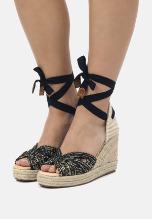 Sandalias de cuña de esparto trenzadas en negro con una suela de cuerda texturizada, que presentan una tira ancha y lazos en el tobillo, acentuadas con detalles en cuero marrón.