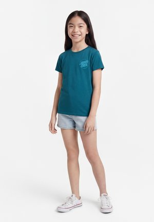 Jong meisje glimlacht, draagt een teal Santa Cruz T-shirt, lichte denim shorts en witte sneakers, staand tegen een effen witte achtergrond.
