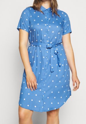 Vrouw draagt een blauw jurkje met korte mouwen, knopen voor, witte polkadots en een ceintuur geknoopt in de taille, staand tegen een effen achtergrond.