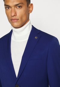 Navyblauer Blazer mit tailliertem Schnitt, kombiniert mit einem weißen Rollkragenpullover. Hat ein einfaches Design und ein kleines Metall-Logo am Revers.