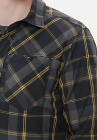 Camicia a scacchi nera e grigia con dettagli gialli, colletto button-down e tasca sul petto con chiusura a bottone. Tessuto testurizzato.