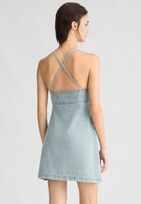 Abito in denim azzurro chiaro con spalline incrociate, forma ad A, orlo grezzo e texture sbiadita; dettagli minimi e design casual.