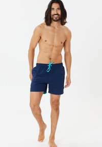 Simshorts gjorda av lättviktsmaterial, marinblå med turkosa snören, knälånga, med en sidficka och en enkel design.