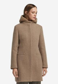 Beige ullkappa med hög krage och foder i päls, med två framfickor och rakt snitt, visad i framvy.