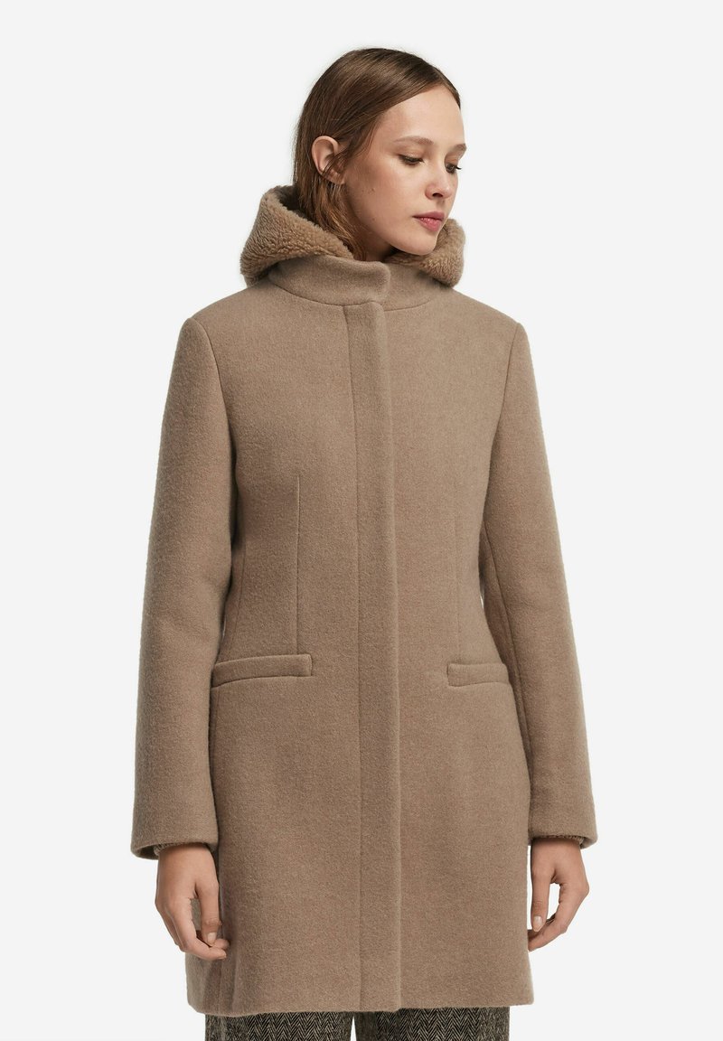 Beige ullkappa med hög krage och foder i päls, med två framfickor och rakt snitt, visad i framvy.