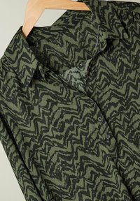 Camisa verde de botones con un patrón en zigzag en verde oscuro y negro. Hecha de una tela texturizada con cuello estructurado y botones.