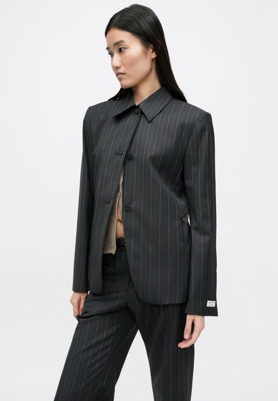 Mustmustrideb kostüümijakk mustade triipudega, kohandatud lõikega, suure kraega, ühe nööbiga sulgemine ning sile tekstuur. Omab peeneid metallilisi aktsente.