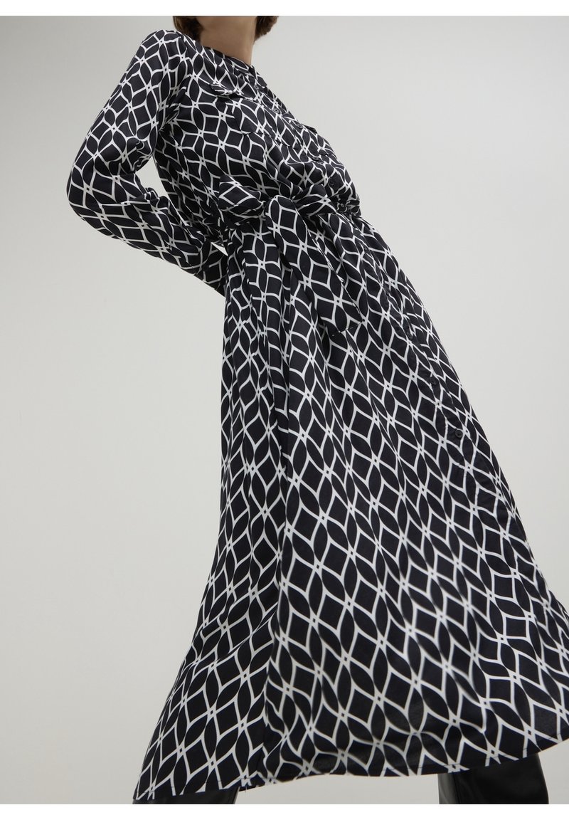 Femme portant une longue robe noire avec un motif en treillis géométrique blanc et une ceinture nouée à la taille, vue en contre-plongée sur un fond uni.