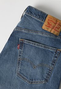 Μπλε τζην Levi's διπλωμένα που δείχνουν πίσω τσέπη με ραφές, κόκκινη ετικέτα Levi's και δερμάτινο λογότυπο με λεπτομέρειες μεγέθους.