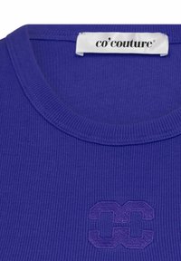 Violet raižytas marškinėliai su apvaliu iškirpimu ir siuvinėtu logotipu, kuriame pavaizduota persipynusi "C" dizaino forma šiek tiek tamsesniame violete.