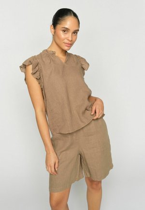 Femme aux cheveux foncés attachés, portant un haut marron sans manches avec des volants et un short assorti jusqu'aux genoux, debout devant un fond clair uni.