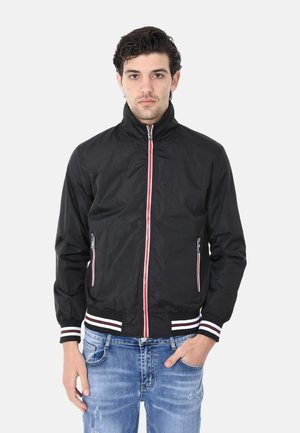 Giubbotto bomber nero con colletto alto, zip frontale, strisce di accento rosse e bianche, e tasche laterali; abbinato a jeans blu strappati.