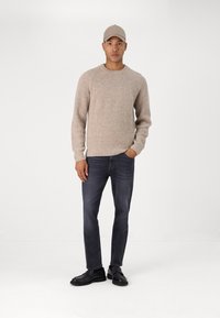 Beige knitted sweater with a textured pattern, paired with dark grey jeans and black shoes, completed by a matching beige cap.