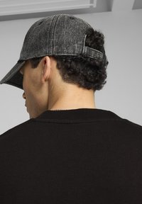 Dark gray cap made of textured material, featuring a curved visor and adjustable strap, worn with a black ribbed sweater.