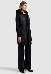 Moteris dėvi ilgą juodą siūtą striukę su gaubtu, juodus plačius kelnes ir juodus aukštakulnius, stovinti prieš pilką foną.