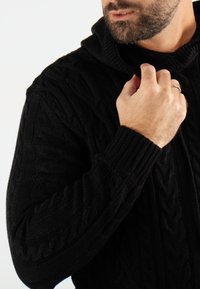 Pull noir en maille torsadée avec capuche. Présente des motifs texturés et des poignets côtelés. Gros plan montrant une main ajustant le col.