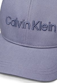 Casquette violette en tissu texturé, ornée d'un logo "Calvin Klein" brodé en bleu marine à l'avant. Forme structurée avec une visière incurvée.