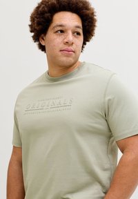 Άτομο με σγουρά μαλλιά και φακίδες που φοράει ένα ανοιχτοπράσινο μπλουζάκι με ανάγλυφο κείμενο "Originals", κοιτάζοντας προς το πλάι μπροστά σε απλό φόντο.