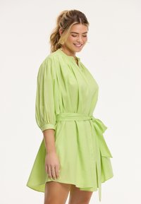 Abito verde chiaro con vestibilità rilassata, abbottonatura frontale e maniche arricciate. Include una cintura in vita legata e un orlo leggermente svasato.