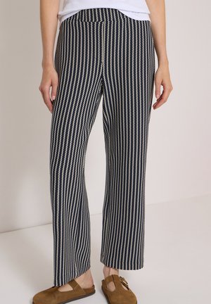 Personne portant un pantalon rayé vertical noir et blanc et des chaussures marron sans lacets avec un haut blanc, debout devant un fond uni.