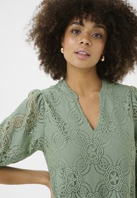 Roheline pitsbluus, millel on lillemustrid, lühikesed puffvarrukad ja V-kaelus, ning detailsemad lõiked kogu kangas.