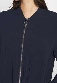 Marinblå idrottsjacka med slät yta, som har en hel dragkedja fram och rund halsringning. Lätt, minimalistisk design.