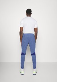Witte T-shirt met korte mouwen, gecombineerd met blauwe sportbroek met een textuurafwerking en contrasterende marineblauwe accenten op de onderbenen. Witte sneakers.